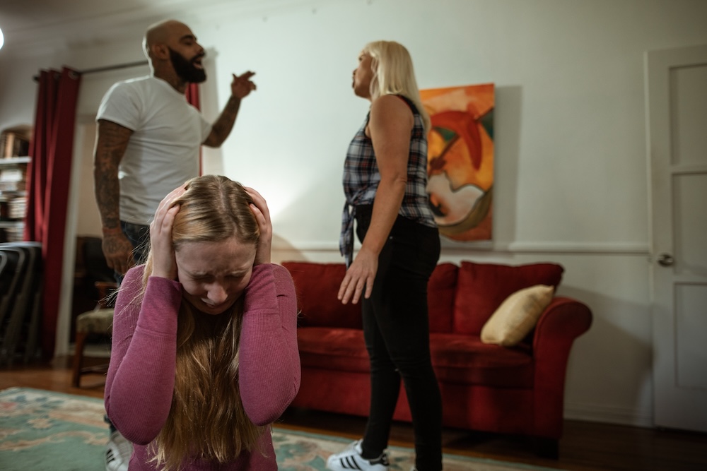 Image of a young girl cowering and blocking her ears while her parents argue in the background, trauma hypnotherapy and ptsd support groups with Rowenna